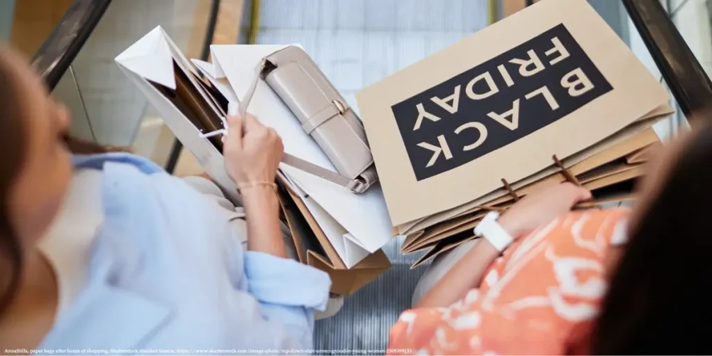 Two women holding shopping bags with "Black Friday" labels on an escalator, symbolizing media consumerism during shopping events.